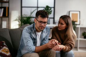 a man with glasses sits on a couch with a woman holding her hand and talking about the signs of iv drug use