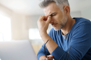 older adult man sits near his laptop looking up long terms effects of alcohol and is distressed and holds the bridge of his nose