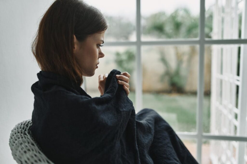 woman wrapped up in a blanket sits in a chair near window and thinks about the effects of heroin