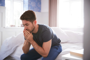 man sits on the edge of bed and leans forward with face resting in his head while thinking about relapse triggers