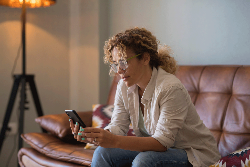 woman with curly hair sits on a couch holding her phone and googling the question does insurance cover rehab