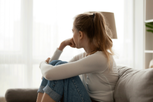 woman sits on couch and looks out nearby window thinking about the connection between trauma and addiction