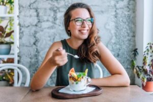 a woman with glasses smiles while eating a bowl of fruit and yoga and learning about benefits of nutritional therapy